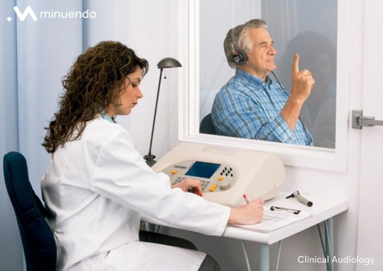 A woman is sitting in front measuring equipment evaluating the hearing of a man behind a glass window.